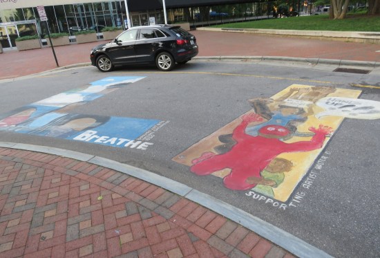 Chalk drawings on asphalt include a red blobby guy with hands up yelling "HANDS UP", and a large blue L that says "I CAN'T BREATHE" along the bottom.