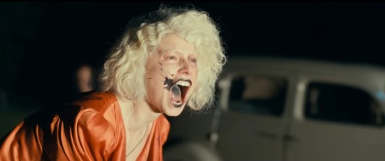 Jessie Buckley with blond hair, black 1930s hat and black smudge next to her mouth, screaming into headlights at night.