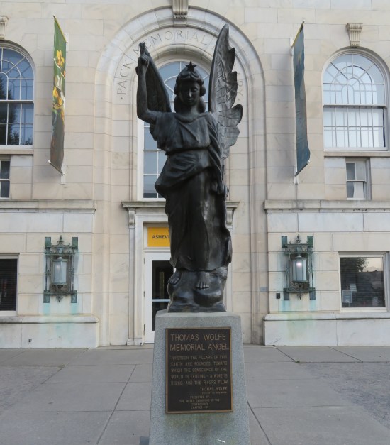 Gray angel sculpture with one hand raised with a peace sign, on a pedestal in front of a two-story formal building.