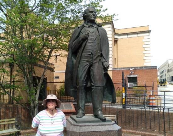 Anne in sun hat posing neutrally next to a tall Andrew Johnson statue. Behind them are a beige brick building and a couple of thin trees.