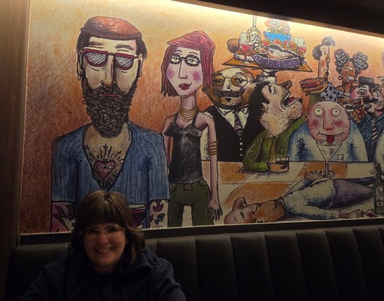 Anne sitting in black restaurant booth On wall above and behind her is cartoon of many partygoers of varying trendiness levels.