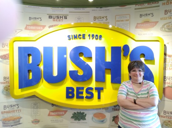 Giant Bush's logo on a wall covered with faded pics of product labels. Anne poses next to it, smiling and arms crossed.