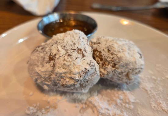 Two round donut holes covered in powered sugar, in front of a tiny metal cup of honey.
