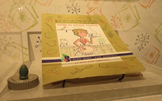 Book called "Bush's Guide to Better Cooking". Front cover image is a 1960s cartoon housewife with an apron, holding a steaming cloche.