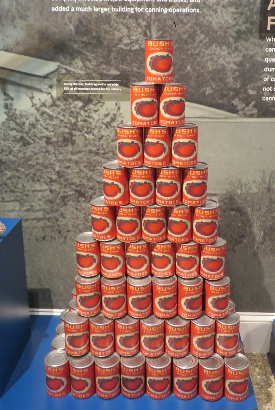 Pyramid of cans of tomatoes. Wall behind them is covered in black-and-white photos.