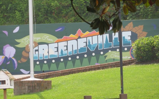Curved green roadside billboard proclaiming in white letters,"Greetings From Greeneville, Tennessee". 