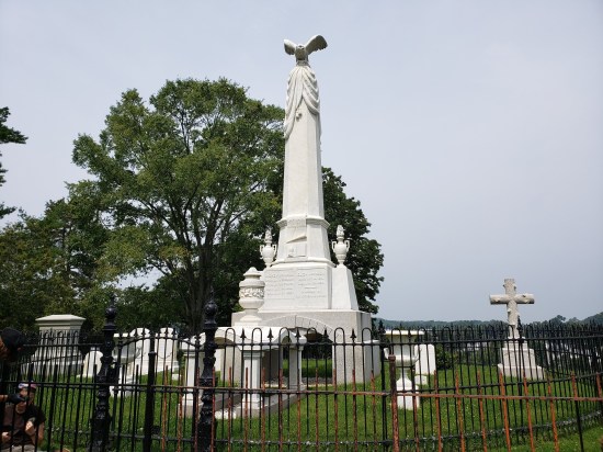 White obelisk and other tombstones behind a black metal fence. Two groundskeepers are working at far left.