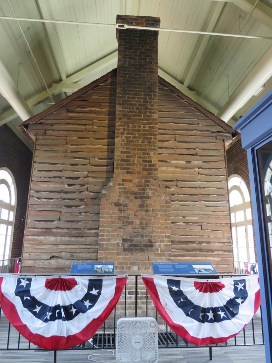 Old wooden building inside a larger newer building, surrounded by a fence with American draperies on it.