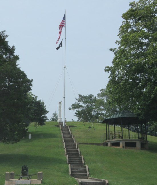 Long staircase up a hill with an obelisk and American flag at the top. A gazebo and two trees are on the hillside.
