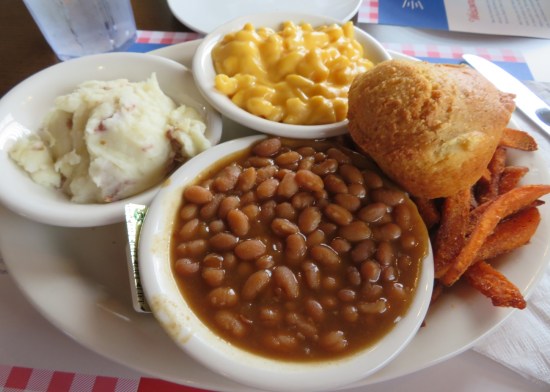Small bowl of baked beans plated with servings of mashed potatoes, corn, sweet potato fries and a roll.0