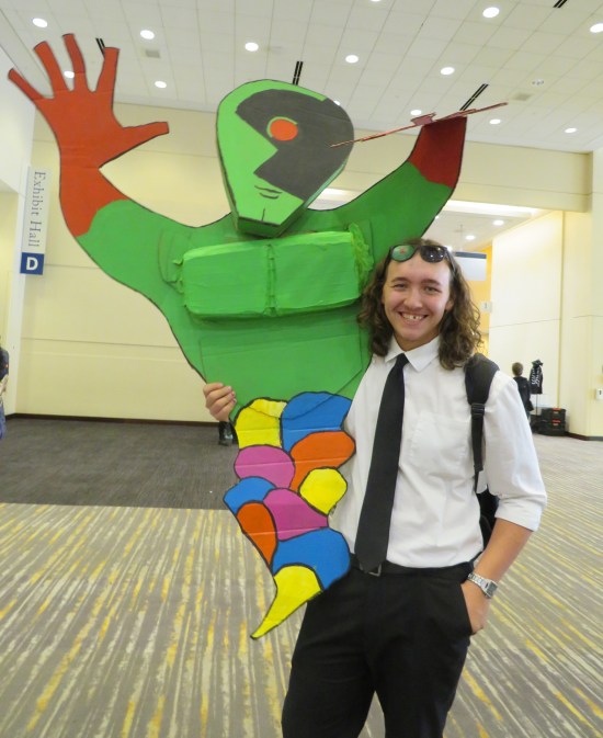 John Jones cosplayer with accurate detective clothing and very specific sunglasses, carrying cardboard replica of psychedelic Absolute Martian Manhunter