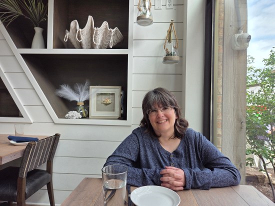 Happy young woman with glasses seated next to a restaurant window. Decor is off-white with light brown flourishes and random white upscale knickknacks.