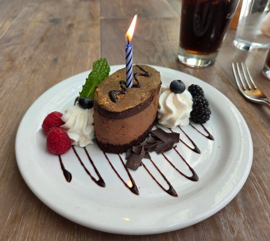 Oval-cylindrical chocolate mousse cake with one lit birthday candle. Also plated are raspberries, blackberries, blueberries, chocolate shavings, whipped cream dollops, and a mint leaf.