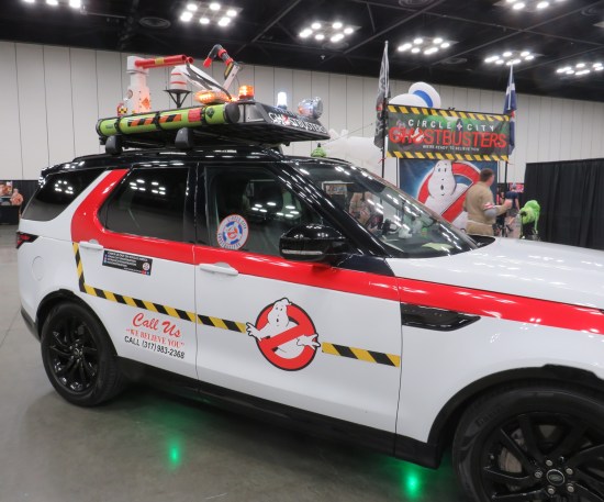 Ghostbusters-themed SUV parked in a comic-con. Circle City Ghostbusters banner is up on poles in background.