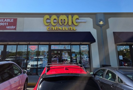 Comic Carnival strip mall storefront with parking spaces all filled. The space next door is for sale.