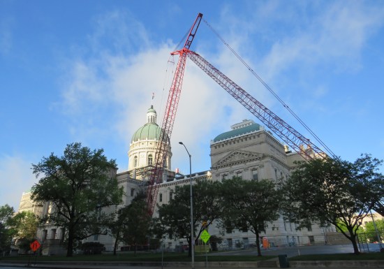 Indiana Statehouse with two red cranes parked in front. Early sunlight strikes a narrow band across them.