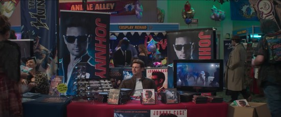 Karl Urban as Johnny Cage manning a comic-con table covered in his own merchandise, mostly awful DVDs.