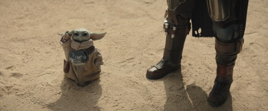 Baby Yoda stands on sand and salutes you. Standing next to him are Din Djarin's shiny boots.