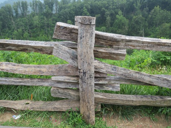 wooden fence intersection in front of forested mountains. Graffiti and carved initials cover the logs.