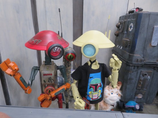 A redheaded pit droid and an offwhite pit droid in a Boba Fett kiddie T-shirt.