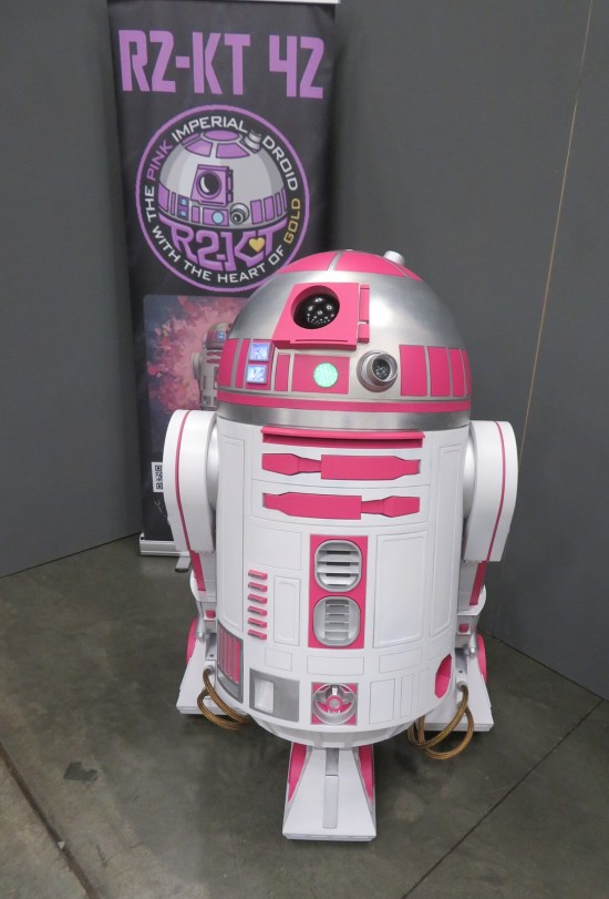 pink astromech droid in front of a banner calling it "The Pink Imperial Droid With a Heart of Gold".