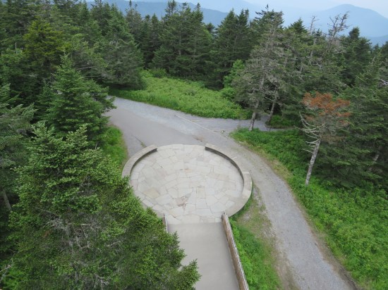 Bird's-eye view of a ramp exit onto a round landing with short wall around it, which in turn leads to a paved forest mountain path.