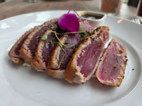several slices of seared ahi stacked like dominoes on a white plate. Topped with light herbs and a purple leaf.