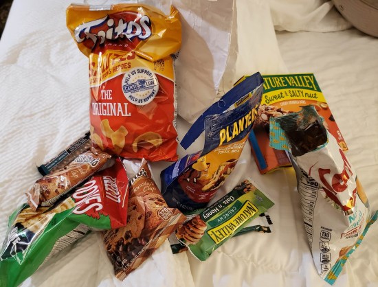 Bags of chips and prepackaged cookies in a pile on a hotel bed.