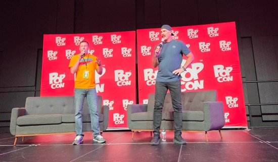 Patrick Warburton on con stage with moderator. Stage has two gray love seats and a red backdrop covered in white Pop Con logos.