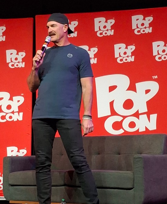 Warburton on stage in gray shirt and backwards cap, black pants and sneakers, and bracelets on his left wrist.
