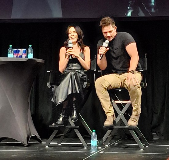 Abigail Spencer and Matt Lanter sitting on stage next to a tall end table holding bottled waters and Red Bull.