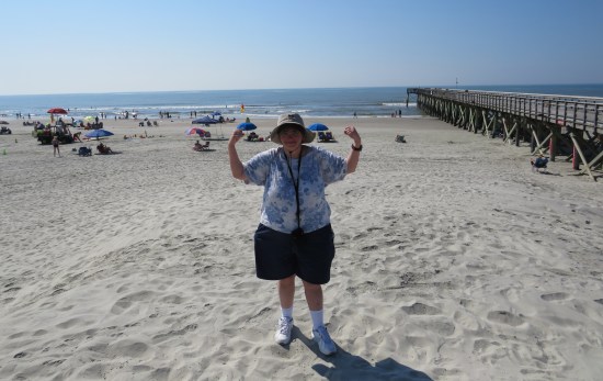 Anne standing on white sandy beach, flexing her biceps. Behind her is an ocean, a pier and sunbathers.