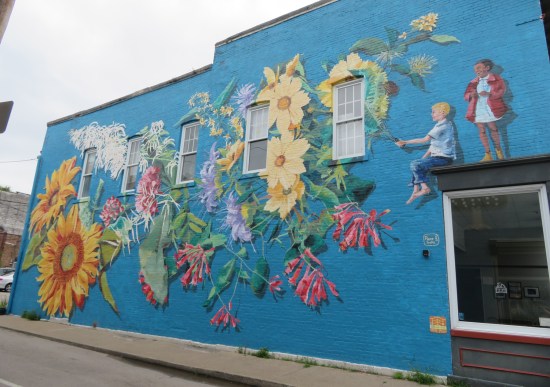 Blue mural with giant bouquet held by tiny boy sitting above real window.