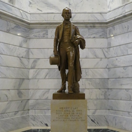 Henry Clay statue in front of a white marble wall, He's holding a top hat, coat, and sheathed sword.