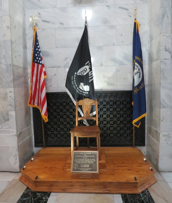 Wooden chair on wooden platform with three flags behind it and a plaque on the front.