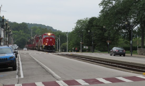 red train coming down tracks that run parallel to the street. Lots of forested hills in the background.