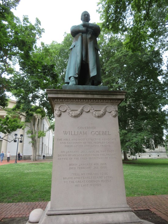 William Goebel statue outdoors on high pedestal, arms crossed and wearing a long coat.
