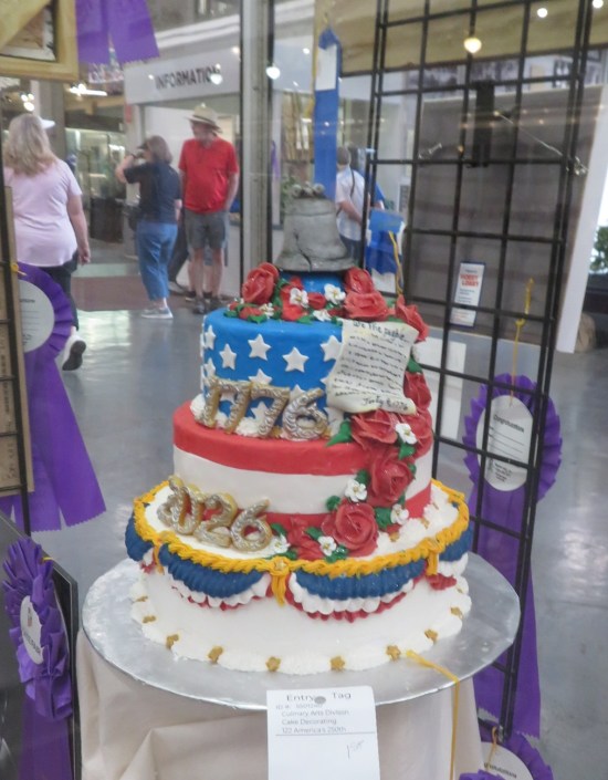 Tall cake in its own vitrine with flag colors, 1776-2026 numbers on the side, and a Liberty Bell topper.