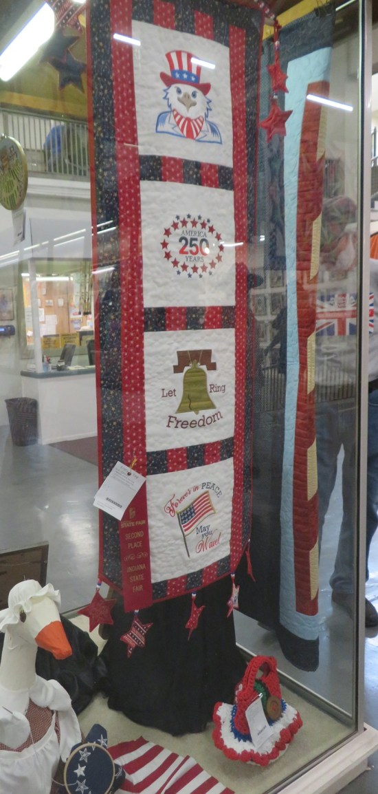 Narrow hanging quilt with pics of a bald eagle with Uncle Sam's hat, the 250th in an oval of stars, a Liberty Bell titled Let Freedom Ring, and a US flag labeled Forever in Peace.