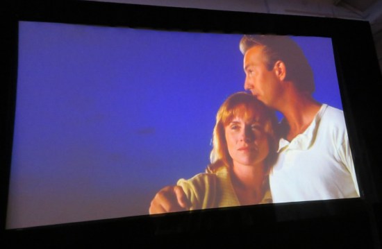 Big TV screen with Amy Madigan and Kevin Costner in old movie, hugging against a blank blue background.