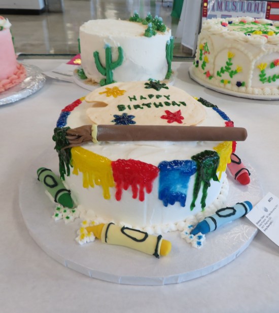 Cake topped with fake dripping paints and paintbrush, and icing crayons around the base.