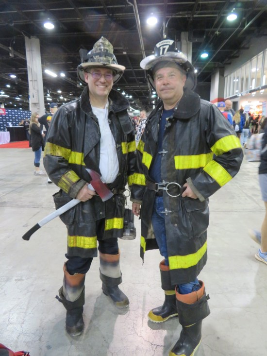 Two guys in firefighter costumes in a comic-con exhibit hall.