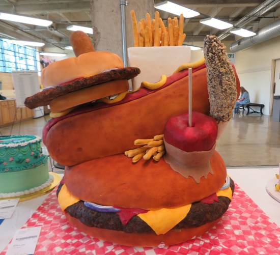 Giant cake shaped like a small burger, fries, hot dog, chocolate ice cream bar, and caramel apple all stacked on one massive burger.