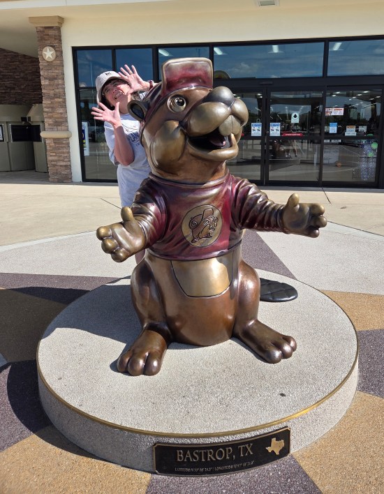 Anne doing jazz hands behind a Buc-ee statue with a Bastrop label.