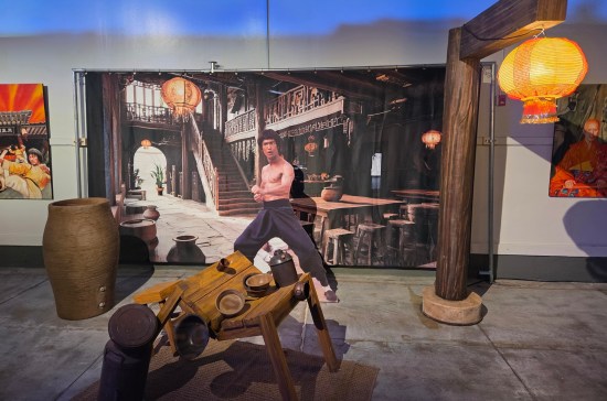 Bruce Lee standee in front of a wall-sized curtain with a kung-fu setting painted on it. East Asian furniture is placed here and there.