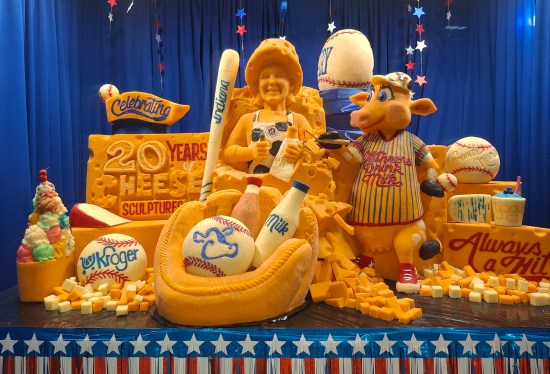 cheese sculpture behind glass in temperature-controlled enclosure. Imagery includes cows, baseball, and self-portrait of the artist.