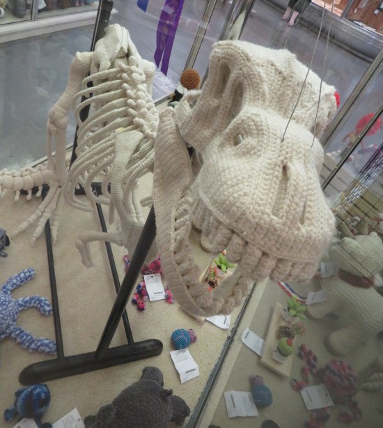 crocheted T-Rex fossil in a glass case looming over other handicrafts.