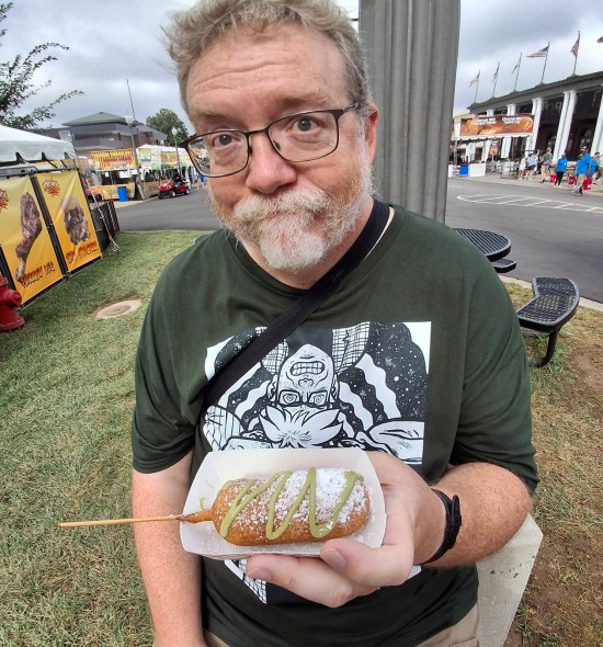 Me holding a snack shaped like a corn dog with green sauce drizzled on it. I seem unimpressed.