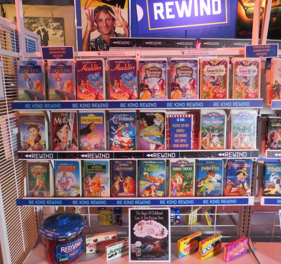 Simulated video rental store with shelves devoted to the 1990s Disney VHS releases in the white cases. Movie candy lines the bottom shelf.