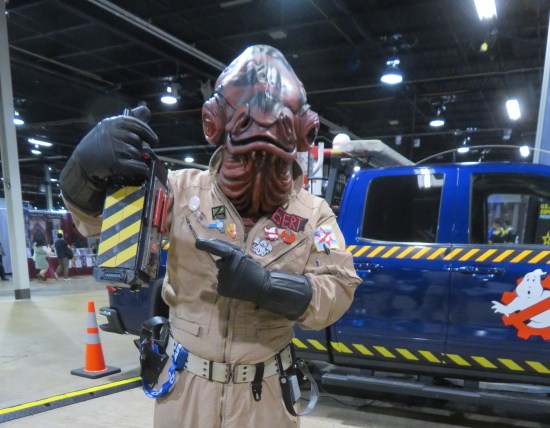 Fan in Ghostbuster jumpsuit and Admiral Ackbar mask holds up a ghost trap.